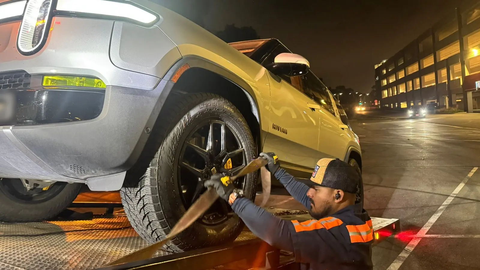 Un conductor de Guerra Towing sujeta un Rivian R1S a una plataforma de remolque por la noche, junto a un estacionamiento de oficinas