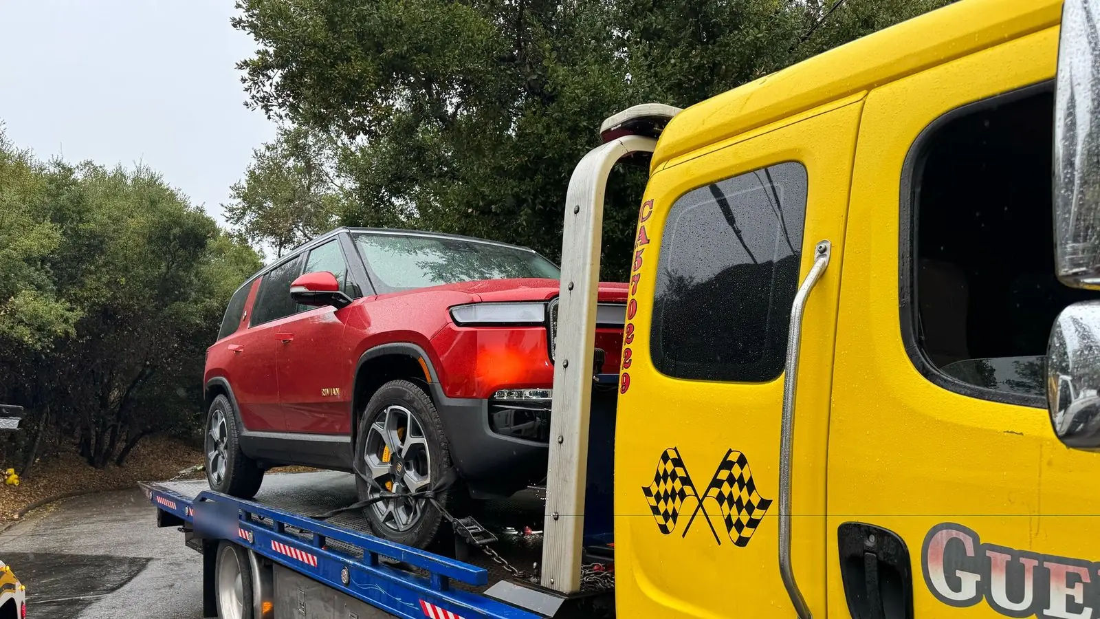 Guerra Towing flat bed carrying a red Rivian electric SUV, strapped down in the rain on a tree-lined foothill street
