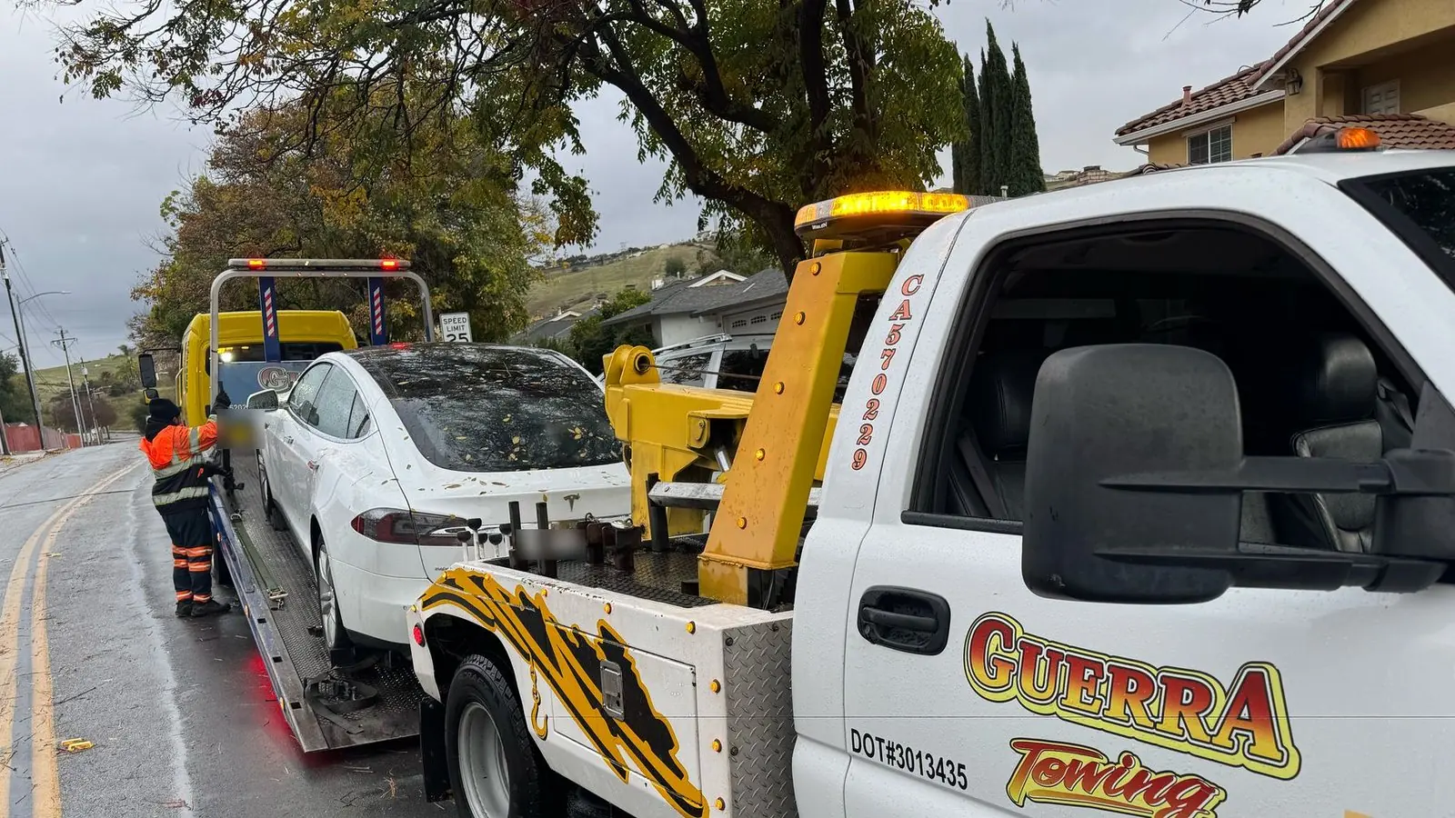 Guerra Towing crew member in a safety vest securing a white Tesla Model S on a flat bed in the rain, with a Guerra wheel-lift truck parked behind